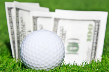 Golf ball on the green with a cup full of hundred dollar bills in the background